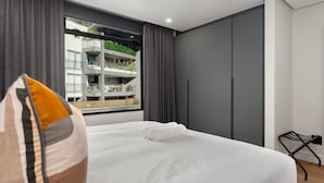 Quarto de hotel moderno com uma cama grande, cortinas cinzentas, vista de um edifício com varandas e uma cadeira preta pequena.