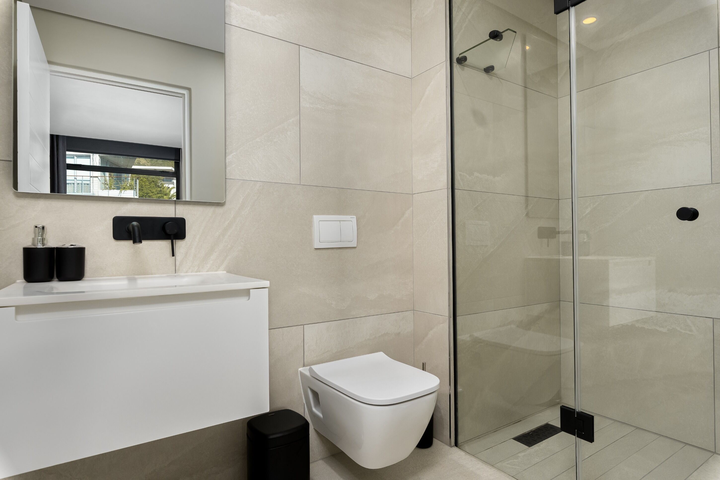 Un baño moderno con cabina de ducha de vidrio, inodoro blanco y lavamanos con dispensadores de jabón negros.