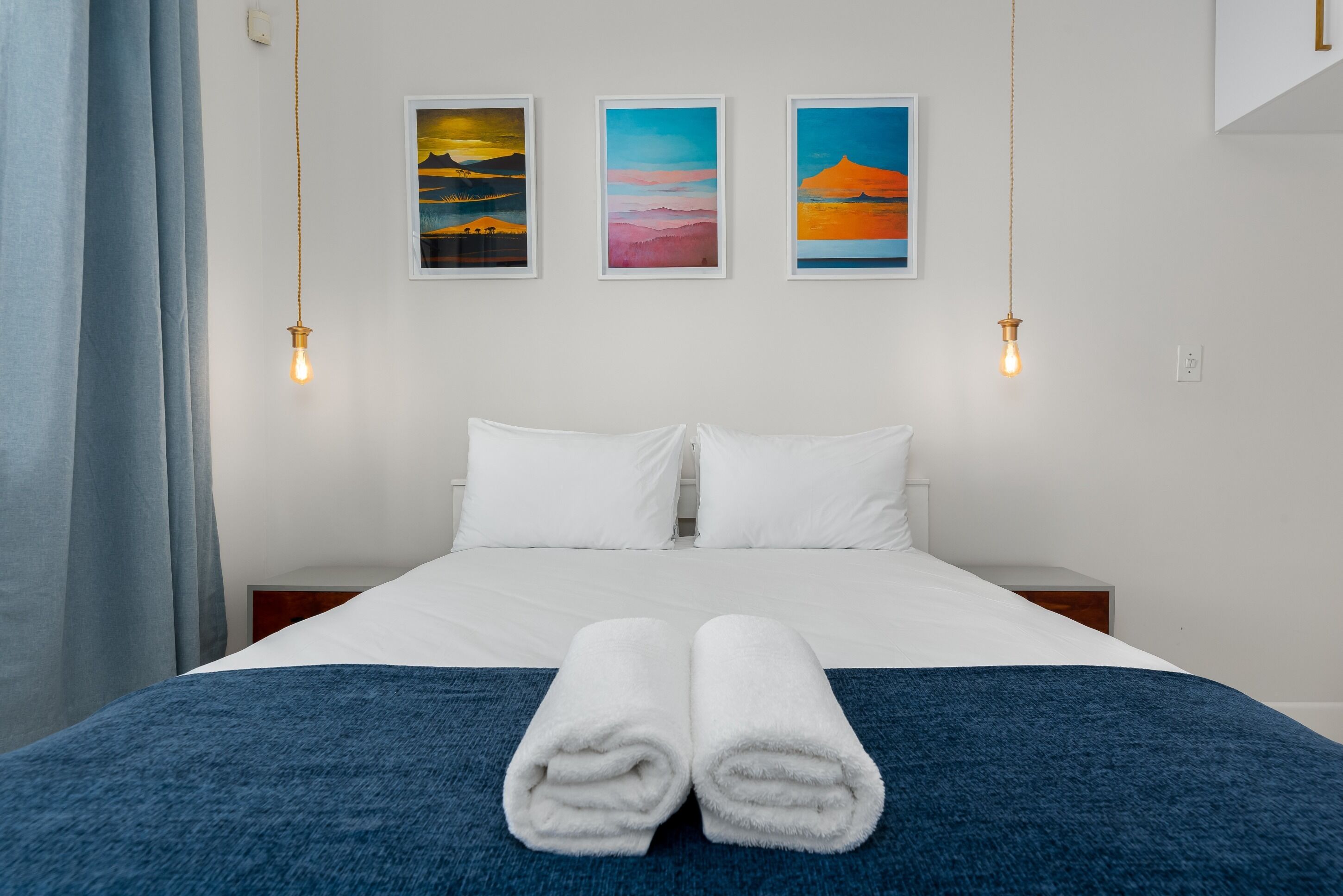 Un lit bien fait, avec des draps blancs et une couverture bleue, deux serviettes blanches posées sur le lit, et trois tableaux encadrés accrochés au mur au-dessus.