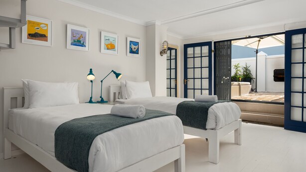 A bedroom with two beds, a nightstand, a lamp, and artwork on the wall. - Grecian Haven In Greenpoint Pool and Braai (Cape Town)