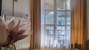 Ein Zimmer mit einem großen Fenster, einem Heizkörper, einem Tisch mit einer Vase und einem grünen Dekorationsgegenstand sowie einem durch das Fenster sichtbaren Stuhl.
