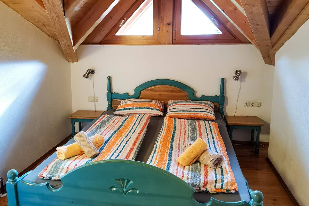 Ein Zimmer mit Holzbalkendecke, geneigtem Dach, einem Bett mit gestreifter Bettwäsche und einem kleinen Tisch.