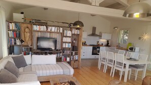 Uma sala de estar acolhedora com sofá, mesa de centro com um tabuleiro de xadrez, estante de livros, televisão e área de cozinha com mesa de jantar e cadeiras.