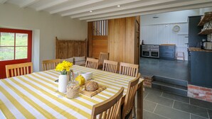 A dining area with a striped tablecloth, wooden chairs, a vase with flowers, a bread basket, and a kitchen visible through an open door. - Pen y Mynydd (Newport)