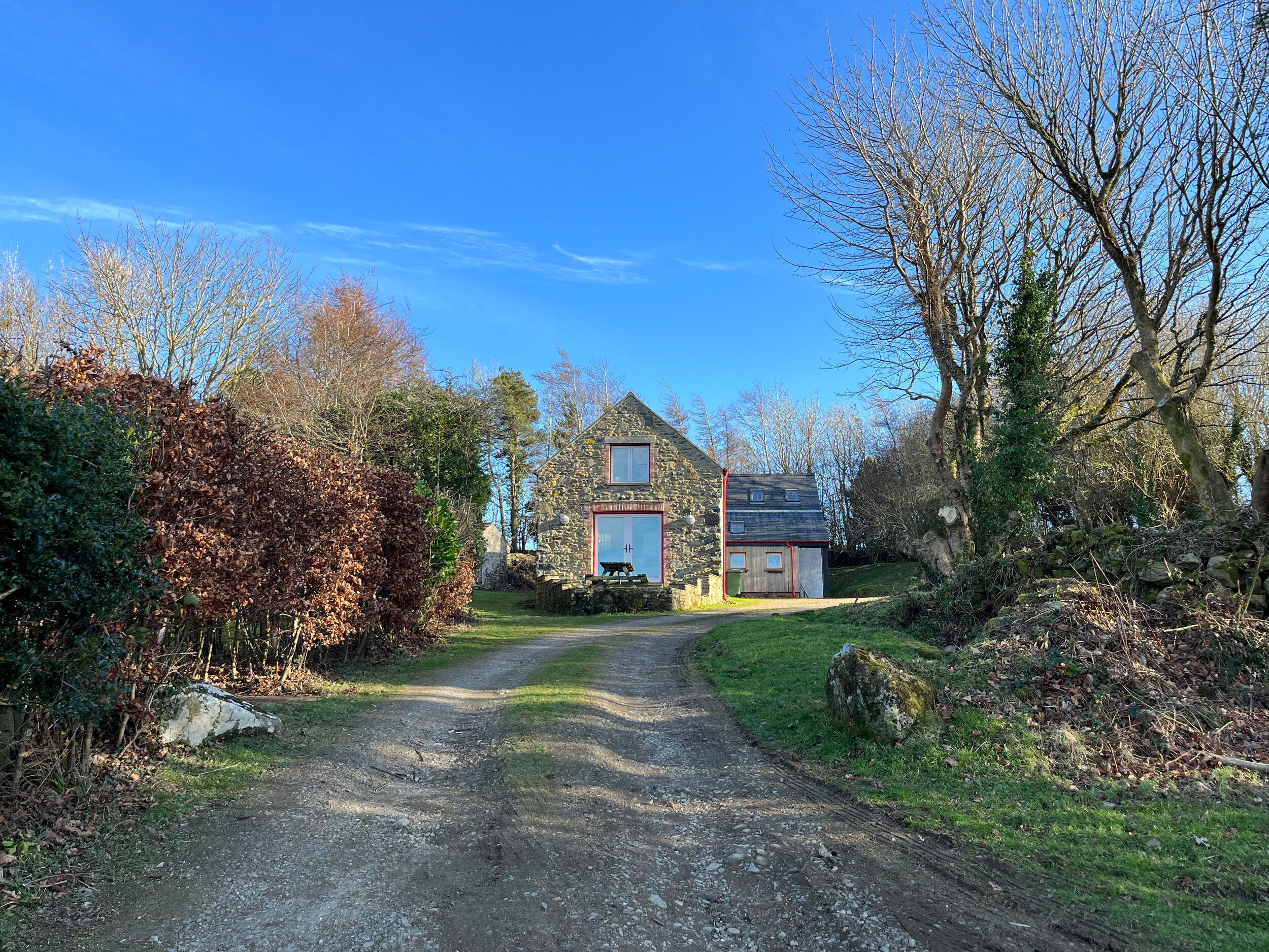 Une maison en pierre avec une allée de gravier menant à la porte d’entrée, entourée d’arbres et d’arbustes.