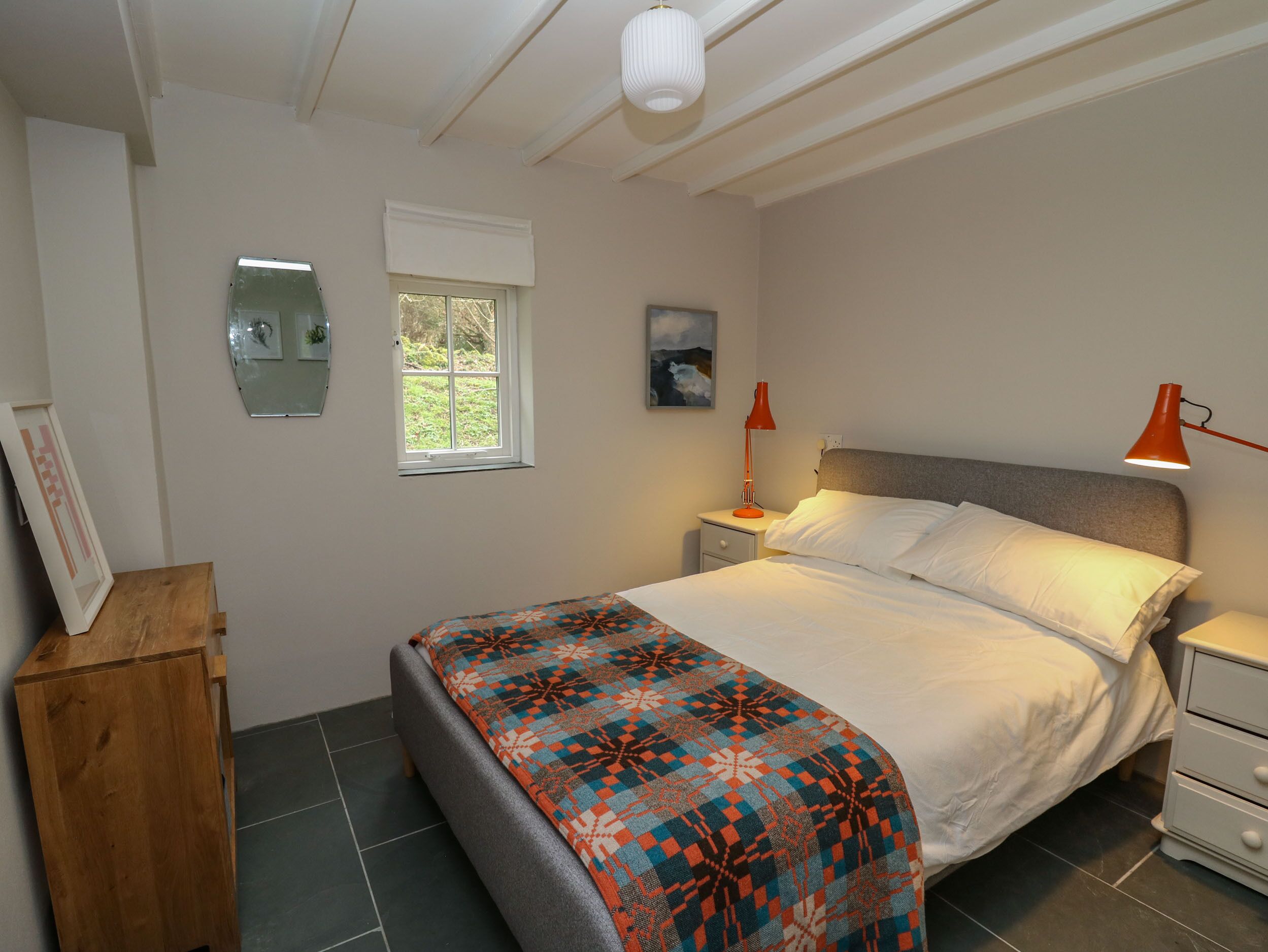 A bedroom with a bed, a nightstand, a wooden cabinet, a window, and a wall-mounted mirror.
