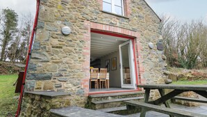 A stone building with a red-framed door and a small window. Inside, there is a dining area with wooden chairs and a table. - Pen y Mynydd (Newport)