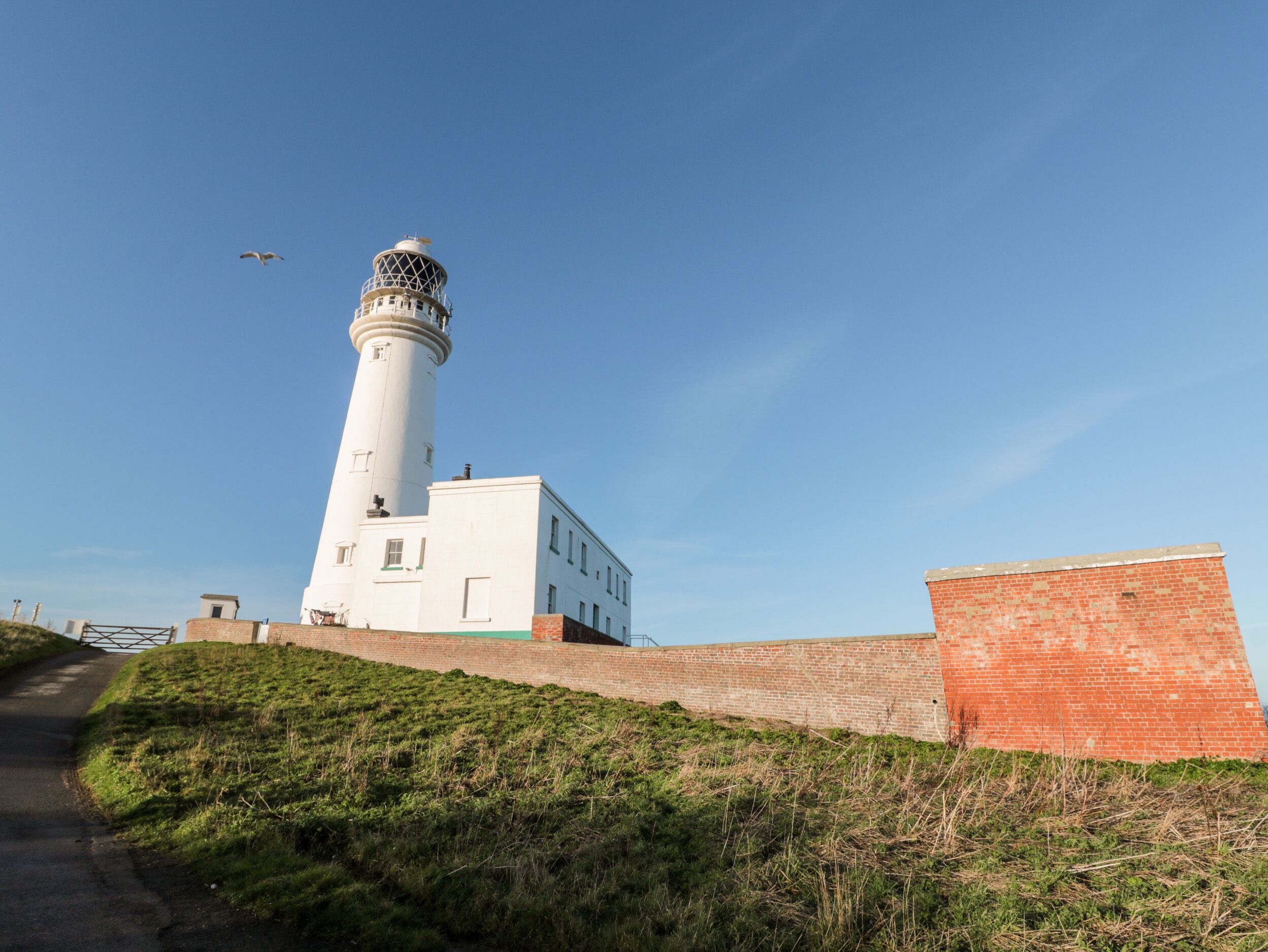 A lighthouse with a white tower and a brick base, surrounded by grassy terrain and a clear sky.
