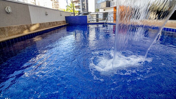 A swimming pool with a water feature, surrounded by a tiled deck and a building facade. - Sun Square Suítes Hotel By Lual (Goiânia)