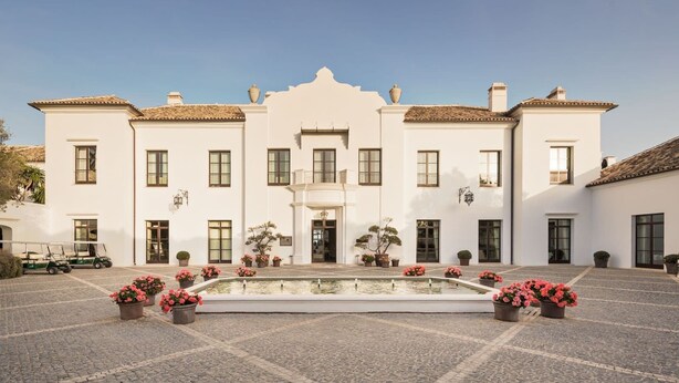 Un edificio grande y blanco con fachada simétrica, entrada central y un patio con fuente y plantas en macetas.