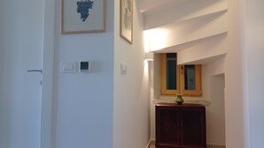 Un couloir avec une porte, un thermostat, une estampe botanique encadrée et un meuble en bois surmonté d’un vase.