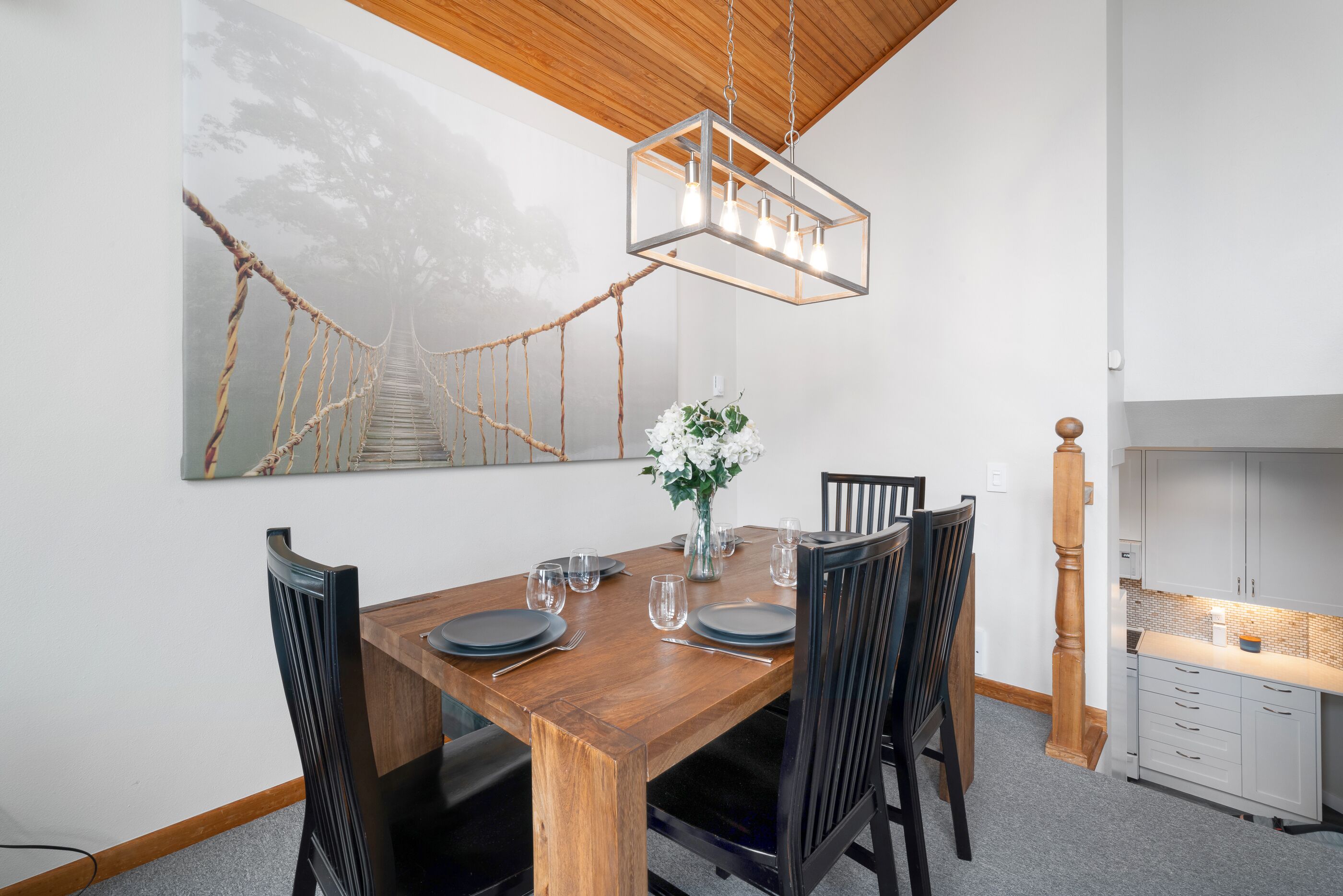 A dining area with a wooden table set for six, surrounded by chairs. A modern pendant light hangs above the table.