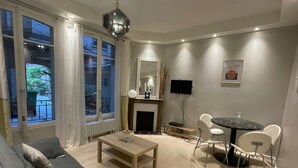 A modern living room with a grey sofa, a wooden coffee table, a fireplace, a TV, and a dining area with white chairs. - Village des Batignolles Paris 17 (Paris)