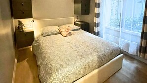 A bedroom with a large bed, a bedside table, a wall-mounted light, and a window with curtains. - Village des Batignolles Paris 17 (Paris)