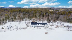 A large, two-story house with a dark roof and white exterior, surrounded by a snowy landscape and dense forest.