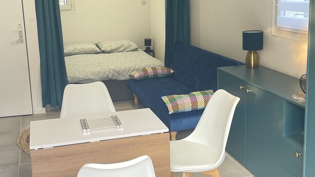 Een compacte slaapkamer met een bed, een bureau met stoelen, een kledingkast en een raam met jaloezieën.