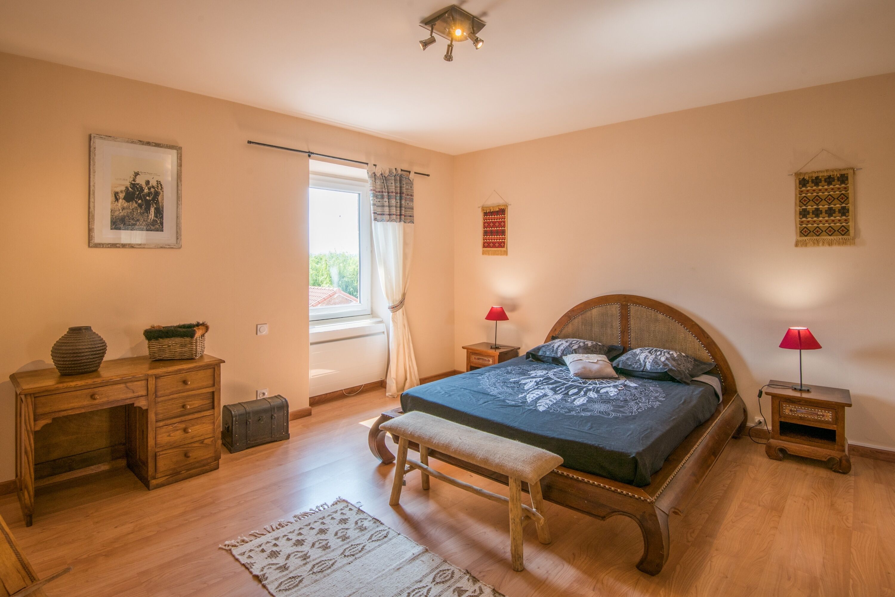 A bedroom with a wooden bed, a desk, a bench, a window with curtains, and a small table with a lamp.