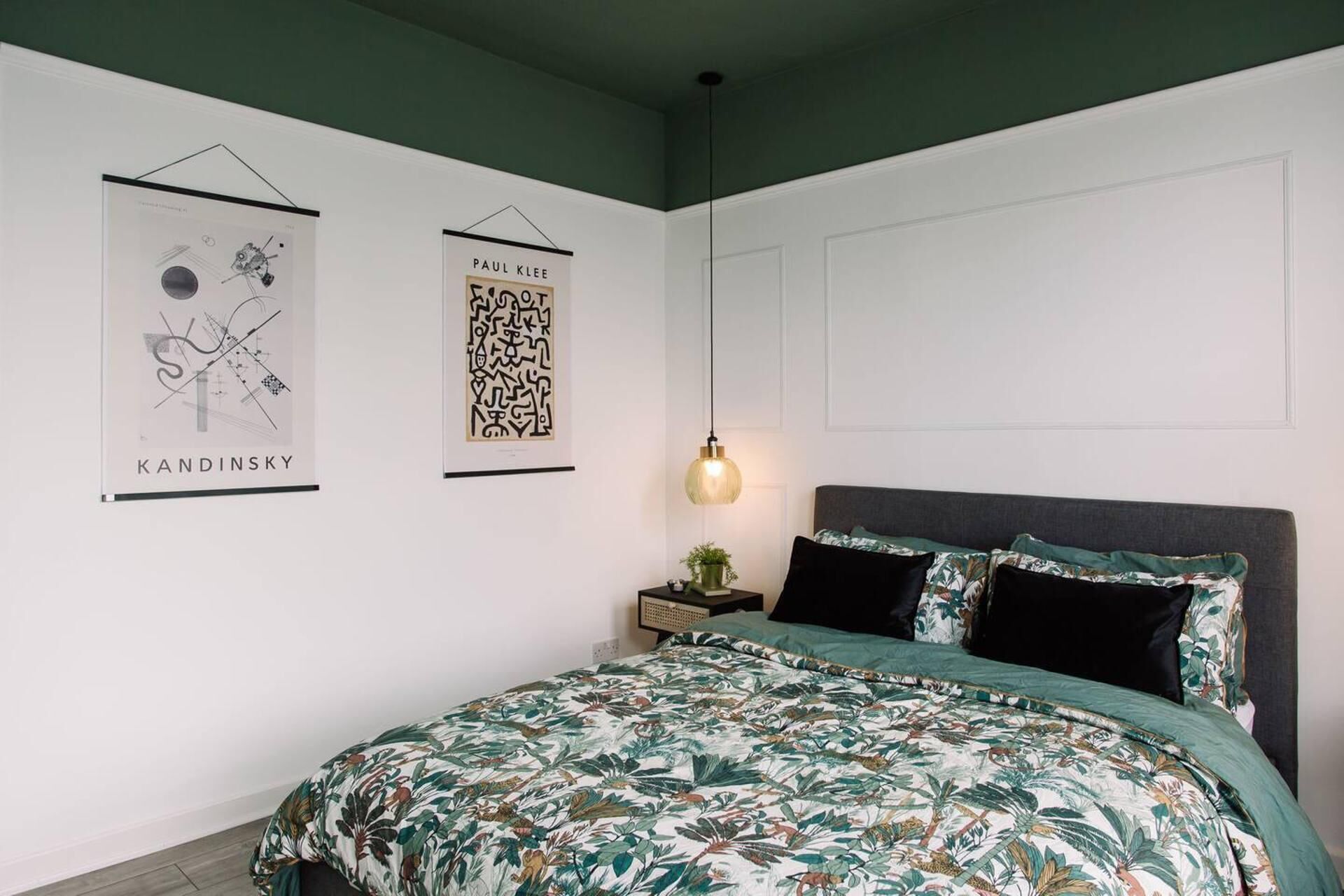 A bedroom with a bed, a nightstand, a hanging light, and two framed artworks on the wall.