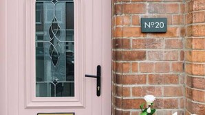 A pink door with a glass panel, a brass mail slot, and a black door handle. A black street lamp is mounted on a brick wall to the right, and a house number sign is visible above it. - Luxury Designer 4-bed Home in the Heart of Belfast (Belfast)