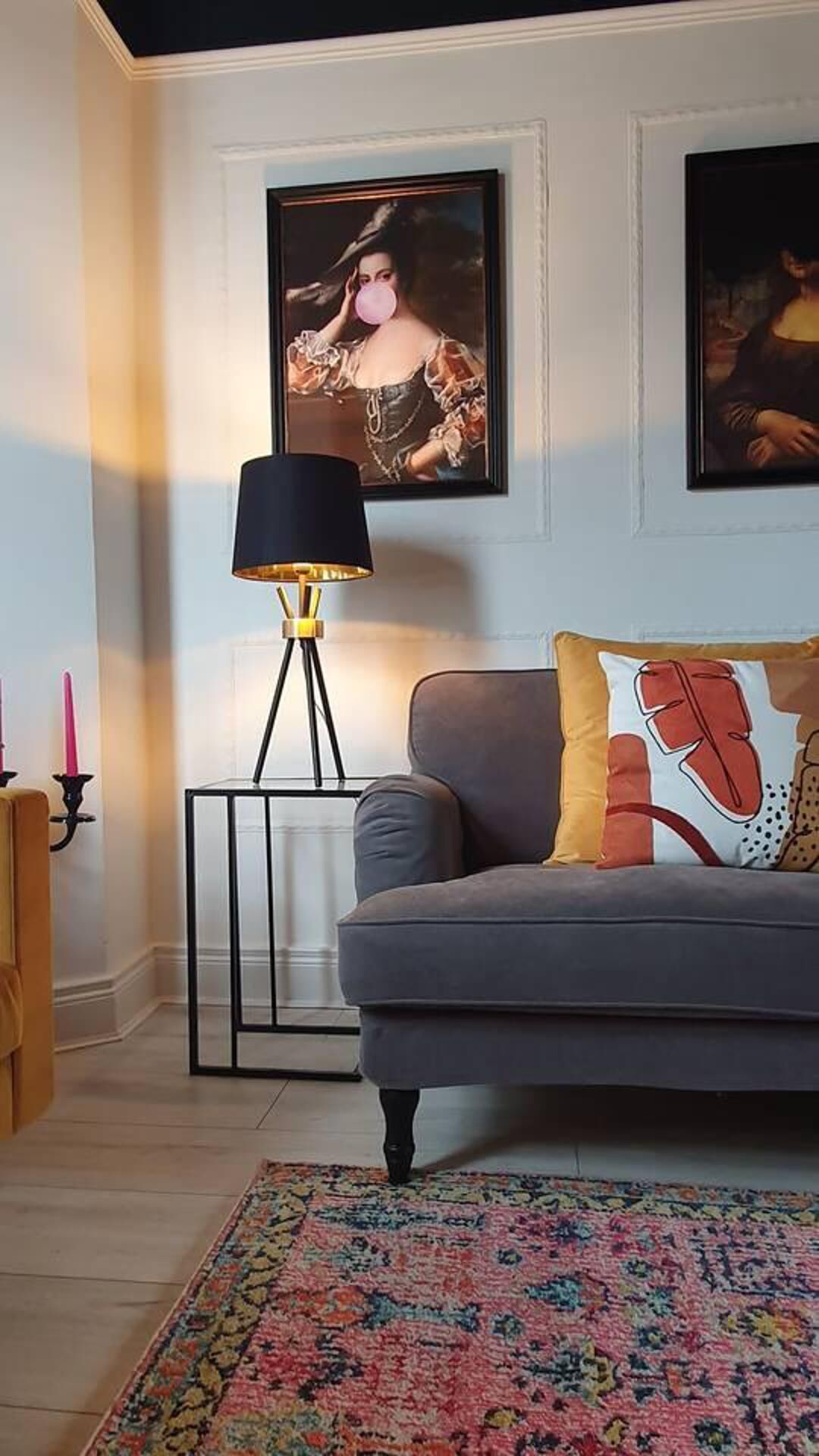 Un salon avec un canapé gris, un tapis à motifs, un lampadaire noir et deux œuvres d’art encadrées accrochées au mur.