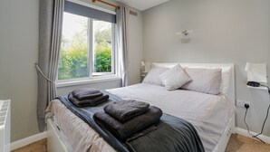 A neatly made bed with a grey blanket and two folded towels, a bedside table with a lamp, and a window with curtains. - Cornish Holiday - 91 Hengar Manor (St Tudy)