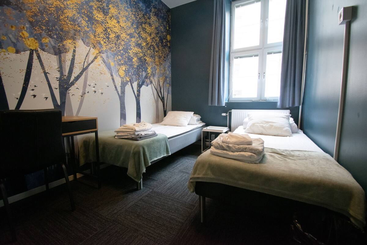 Quarto de hotel com duas camas de solteiro, uma escrivaninha, uma cadeira, uma janela com cortinas e um mural na parede.