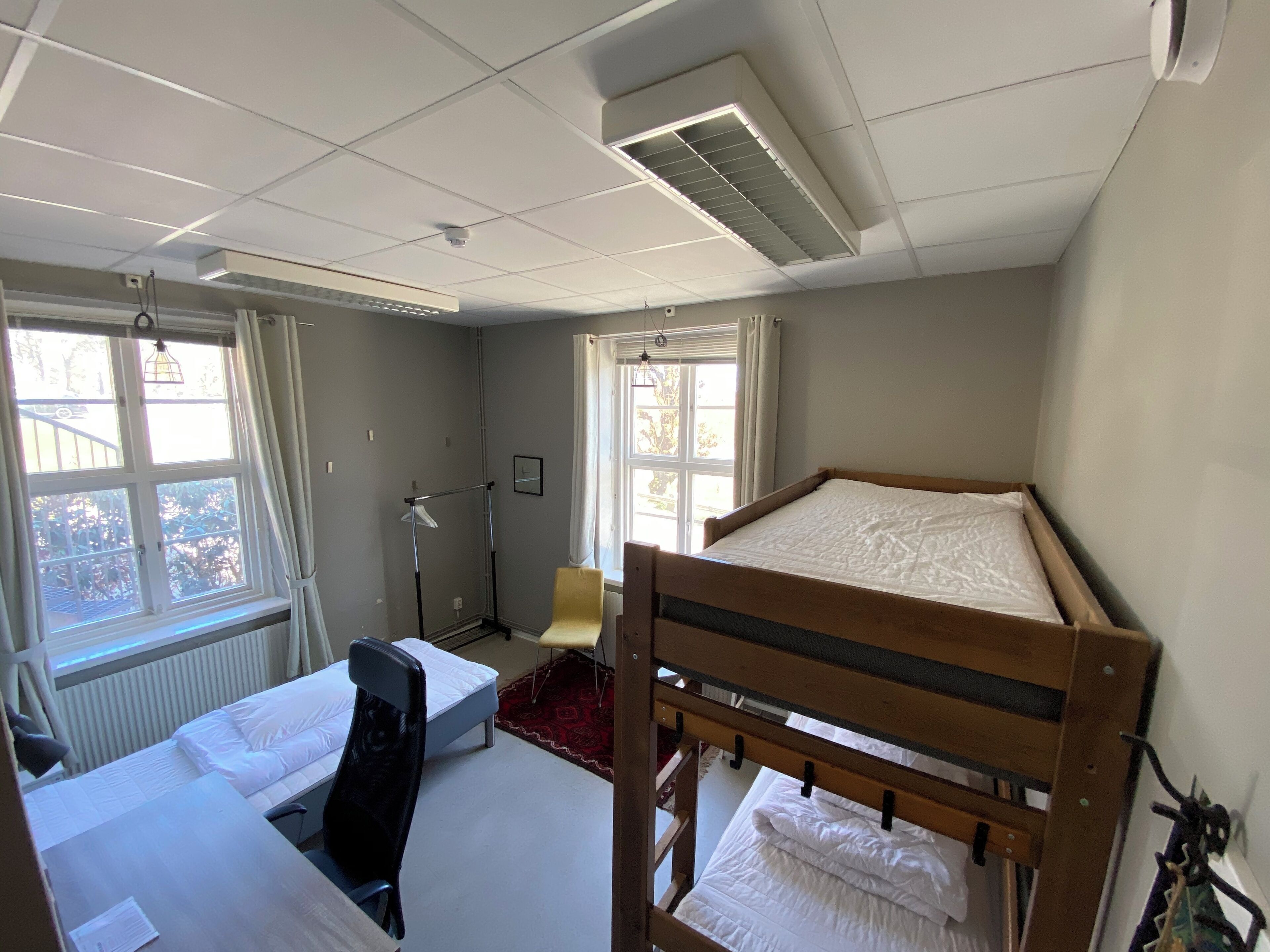 Ett studentrum med våningssängar, ett skrivbord, en stol, ett element och ett fönster med gardiner.