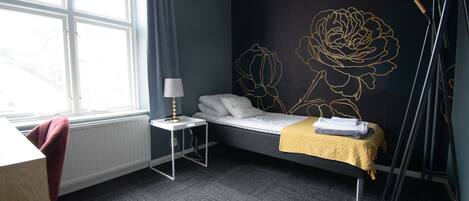 A room with a bed, a small table with a lamp, a chair, and a window.