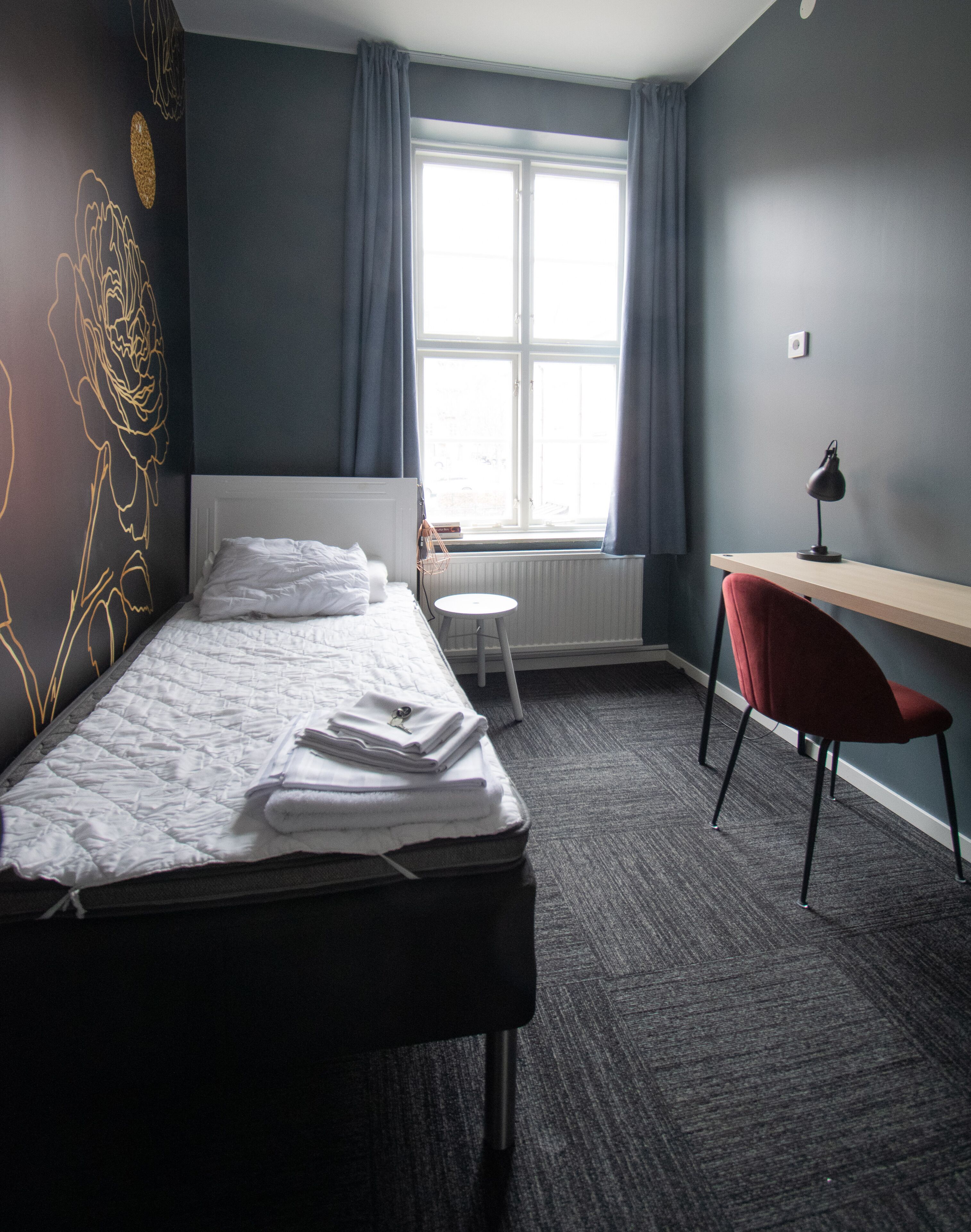 Een hotelkamer met een bed, een klein rond tafeltje, een bureau met een stoel en een raam met gordijnen.
