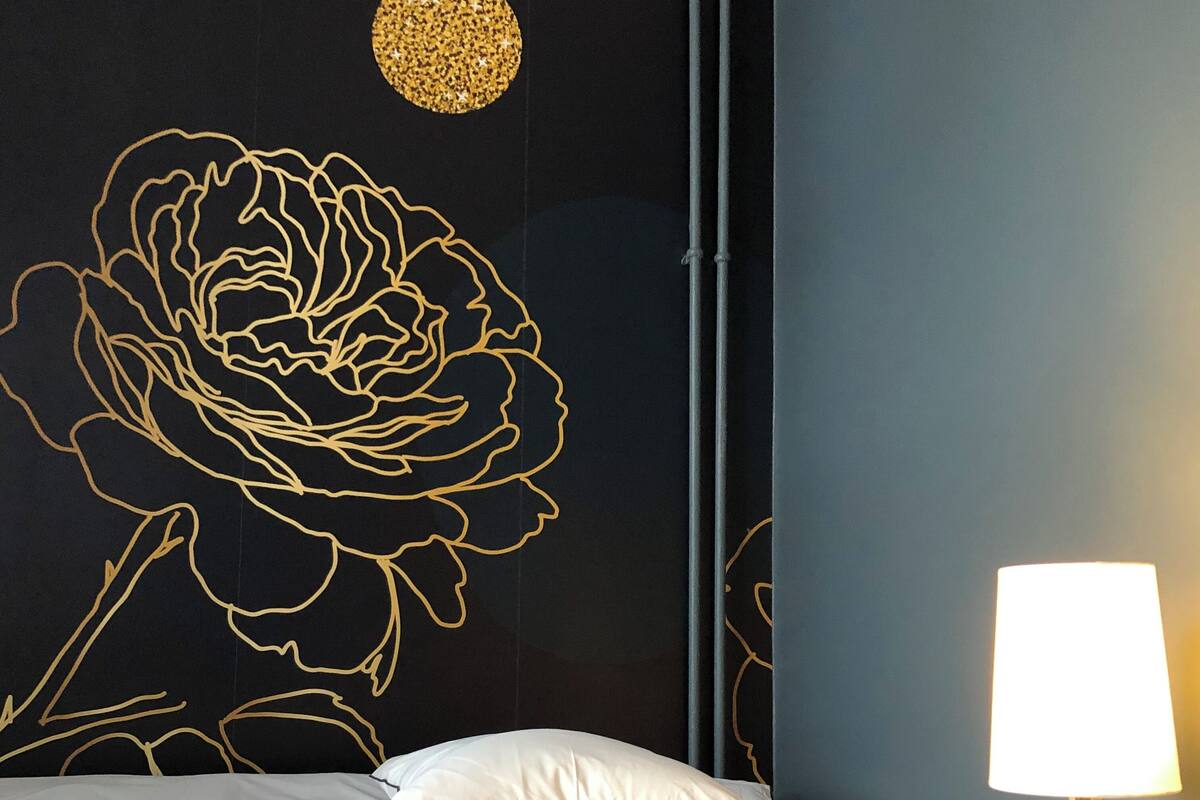 Quarto com parede preta adornada por um grande desenho floral dourado, cama branca com duas almofadas e mesa de cabeceira com abajur.