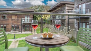 A wooden table with two glasses of red wine and two cupcakes on a balcony with a view of a wooden building and a grassy area.