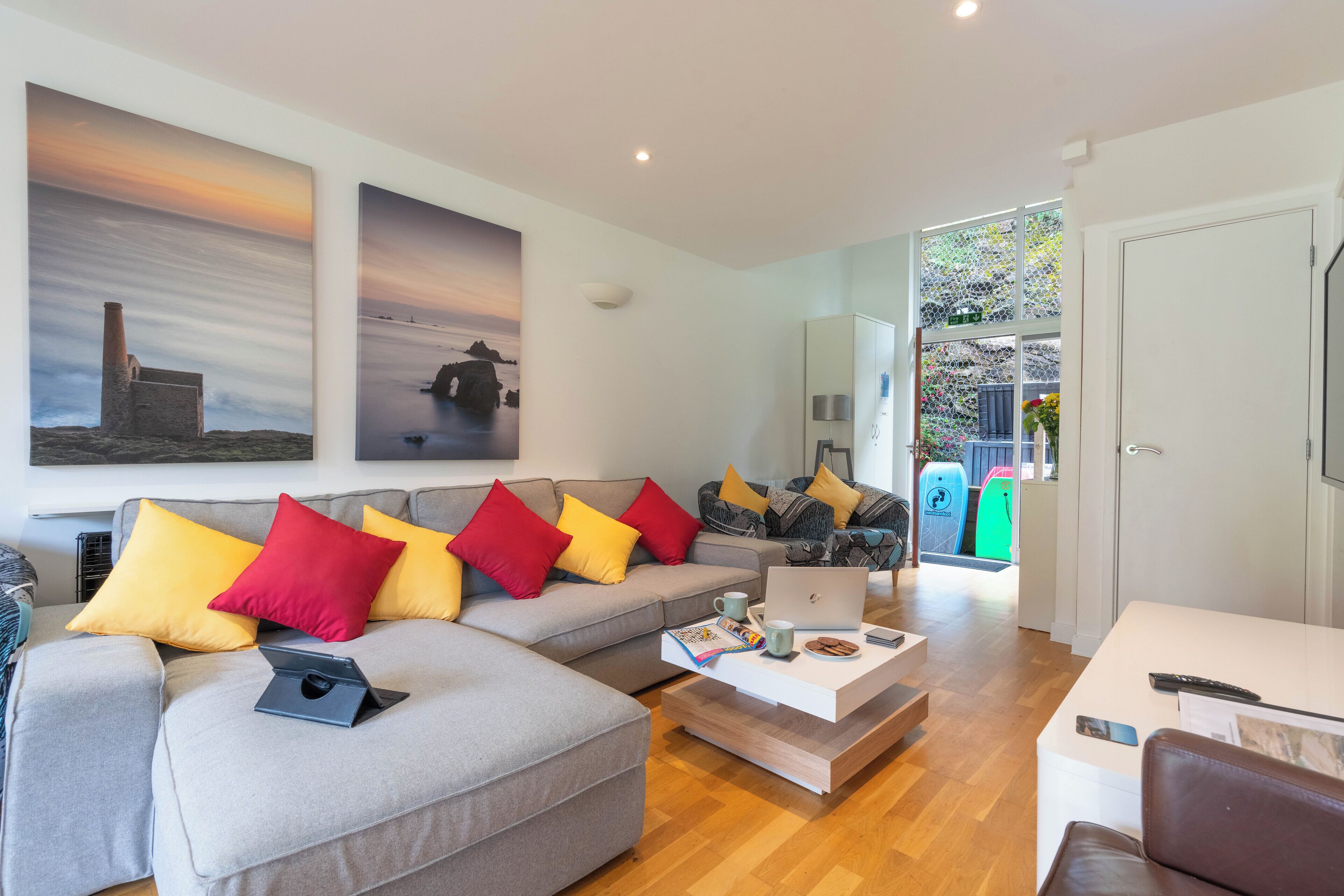 A modern living room with a grey sofa, colorful cushions, a coffee table with a laptop, and a large window with a view of the sea.