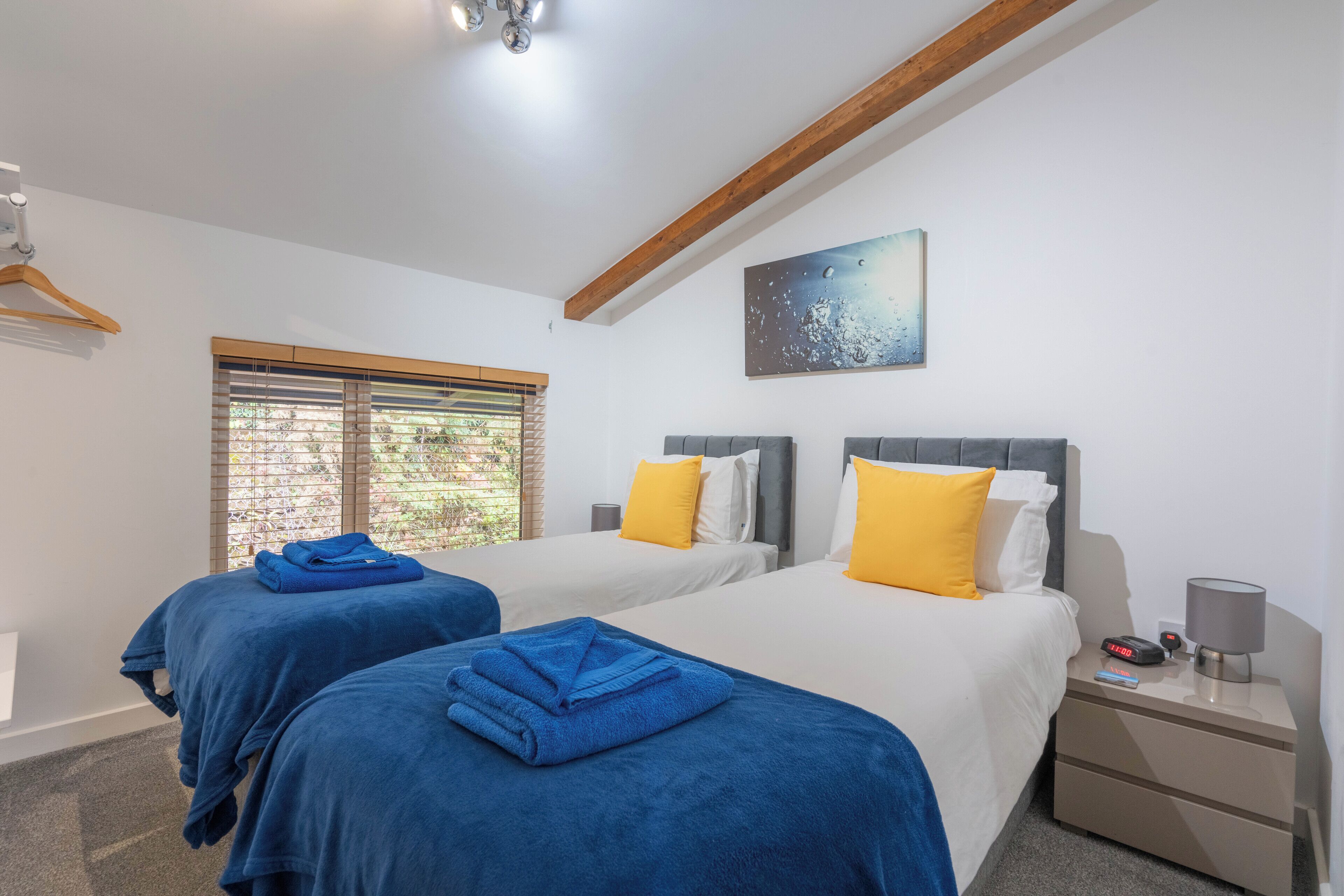 A hotel room with two beds, each with blue and white bedding, a painting on the wall, and a window with blinds.