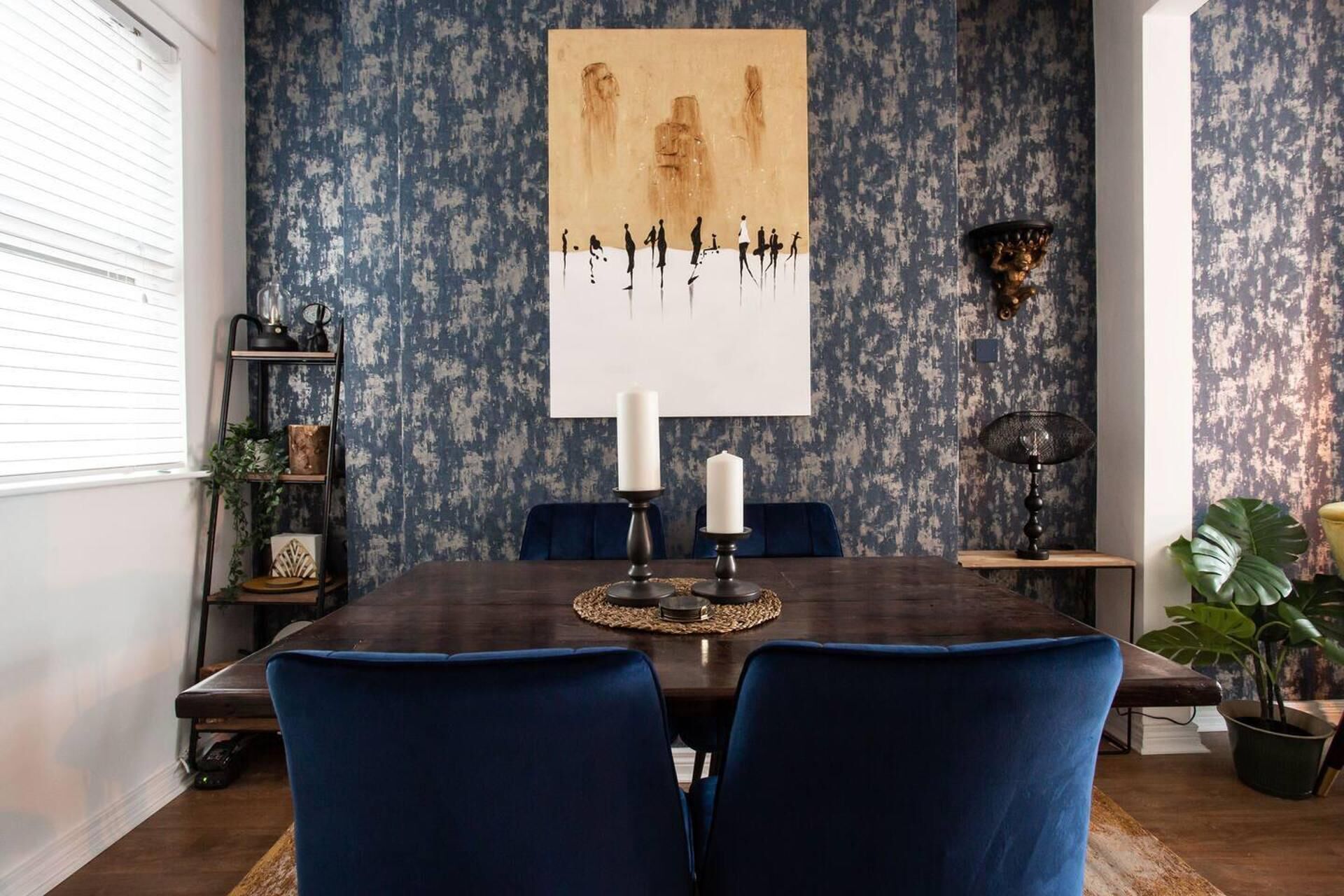 A dining area with a dark wooden table, two blue chairs, a white wall with a large abstract painting, and a blue patterned wallpaper.