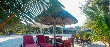 Un salón junto a la playa con sillas reclinables rojas y una mesa bajo un toldo de paja.