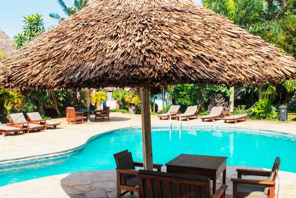 A pool area with a thatched umbrella, lounge chairs, and a stone-tiled deck.