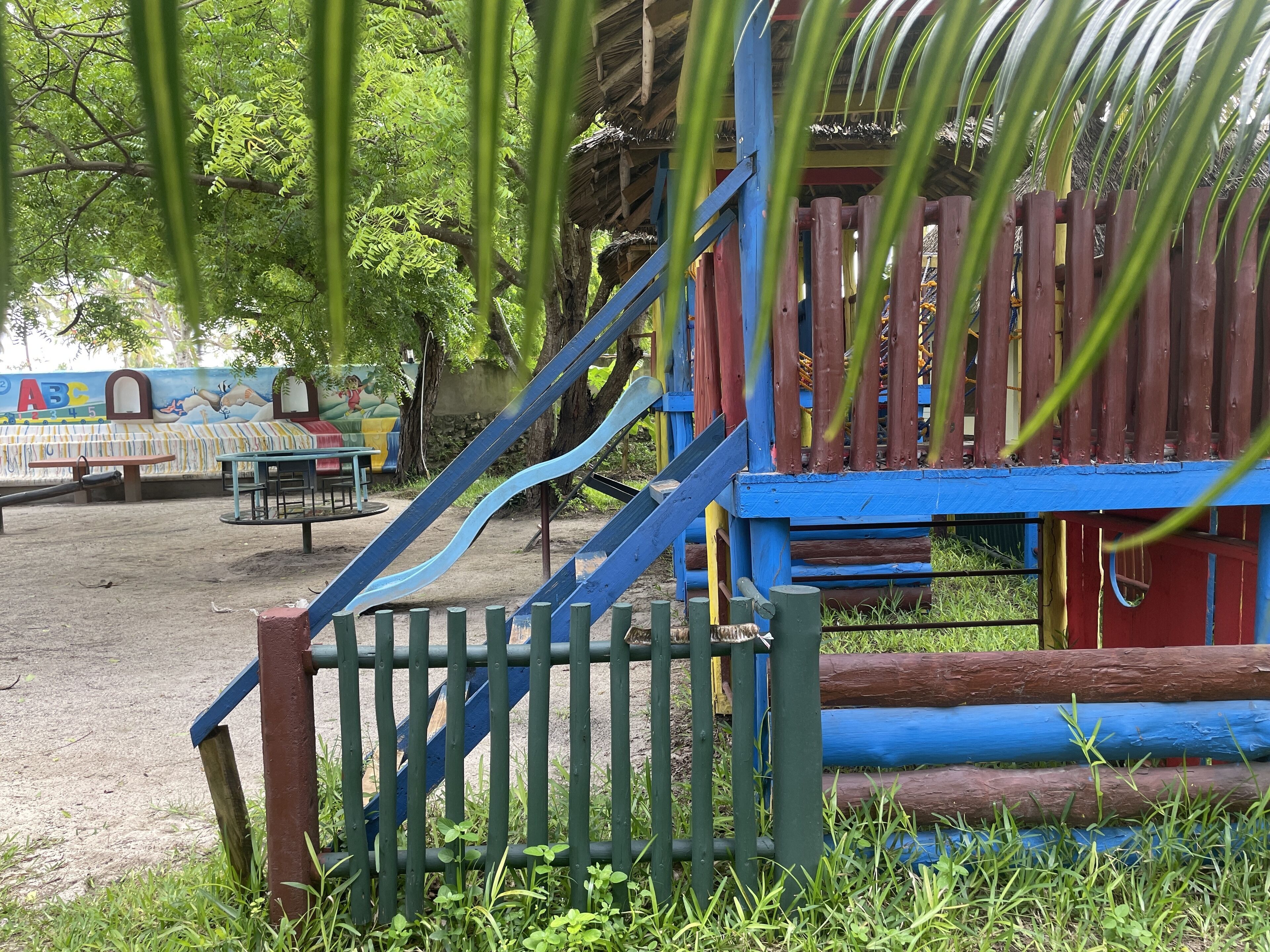 A playground with a slide and climbing structures, surrounded by trees and a mural on the wall.