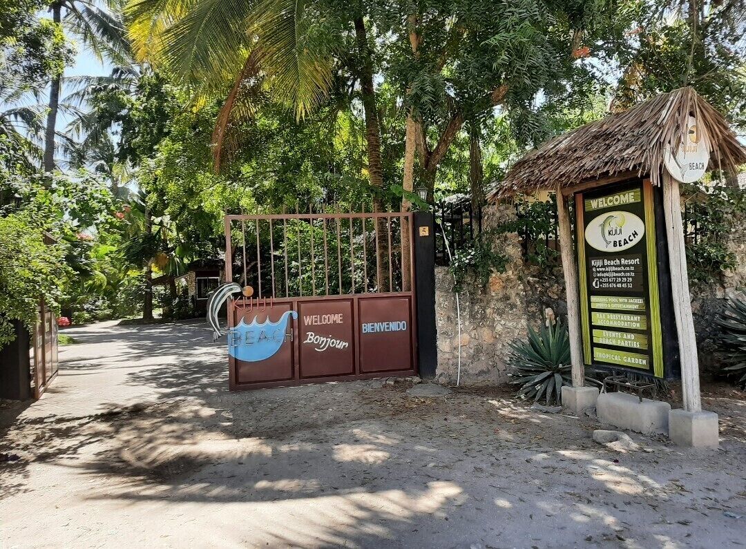 Entree van een tropisch resort met een bord dat bezoekers verwelkomt bij Kijii Beach Resort, met een tropische tuin en een constructie met rieten dak.