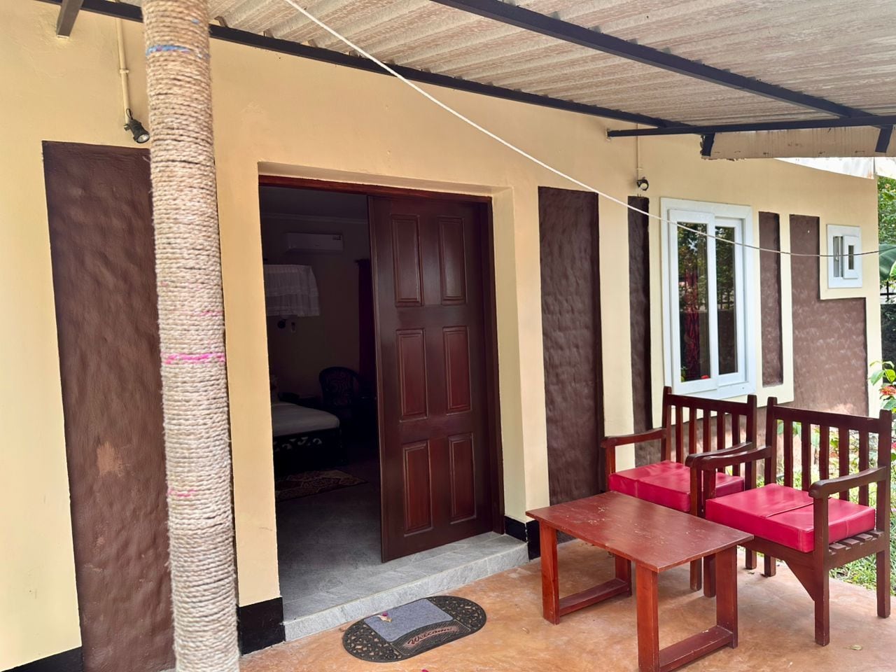 A covered porch with a wooden table and chairs, a door leading to an interior room, and a small outdoor area with a mat.