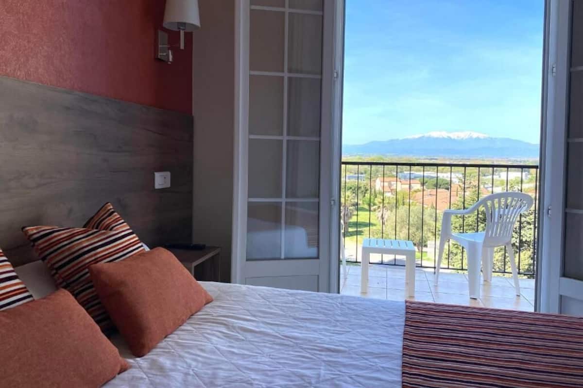 Habitación de hotel con cama, mesita de noche, lámpara y vistas a un balcón con sillas.
