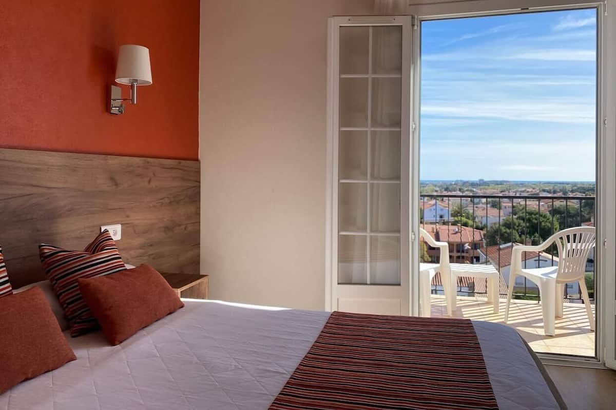 Un dormitorio con una cama grande, vistas de la ciudad a través de un balcón y una pared blanca con una lámpara.