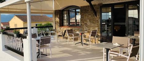 Un patio exterior con muros de piedra, varias mesas y sillas, y una zona de estar techada.
