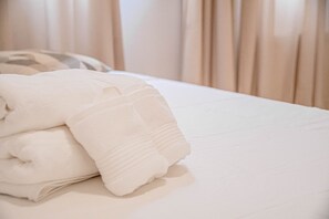 Una toalla blanca doblada con cuidado sobre una cama con una colcha blanca lisa y cortinas color beige al fondo.