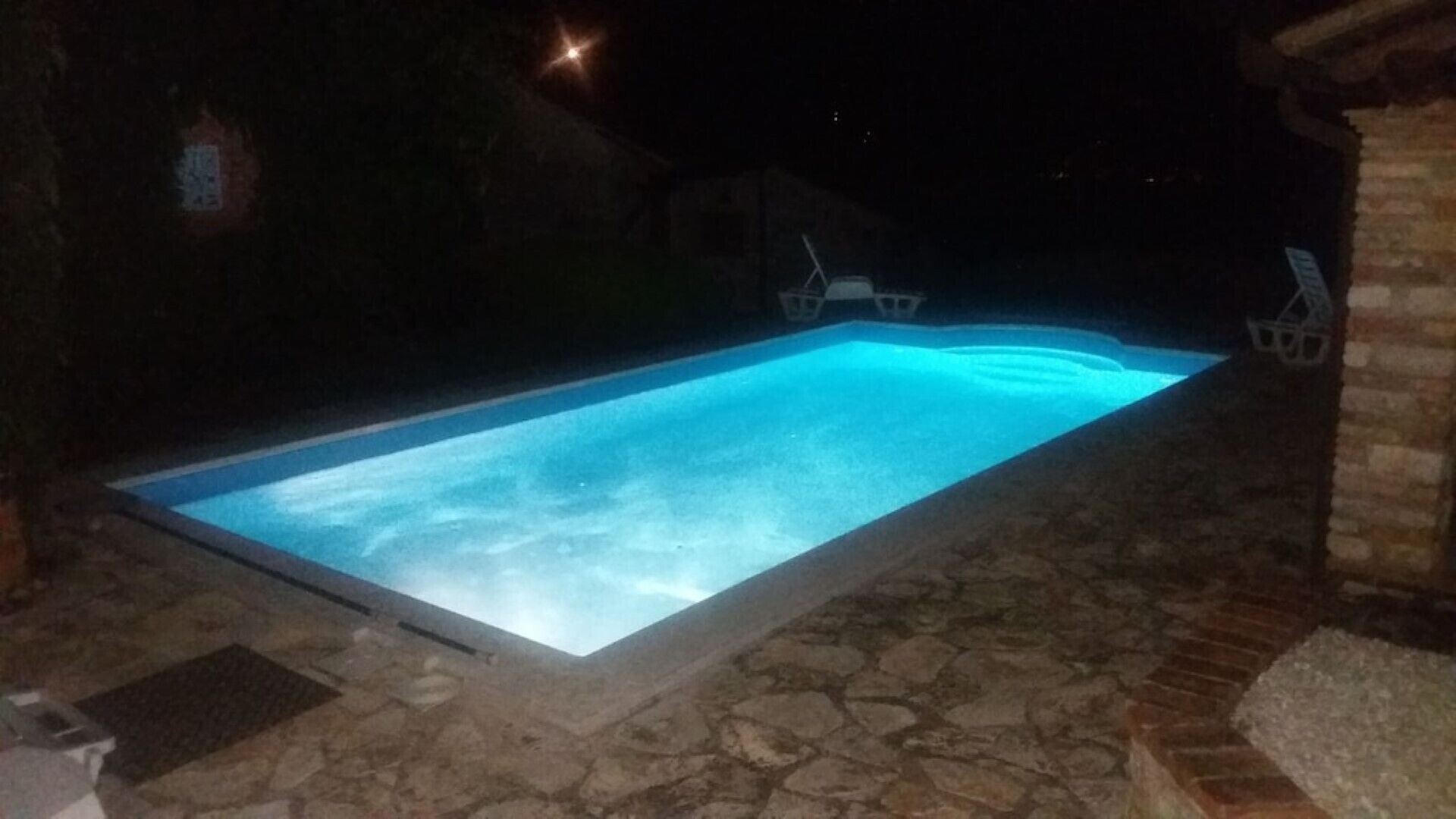 A rectangular swimming pool with a clear blue water surface, surrounded by stone tiles and a brick structure.