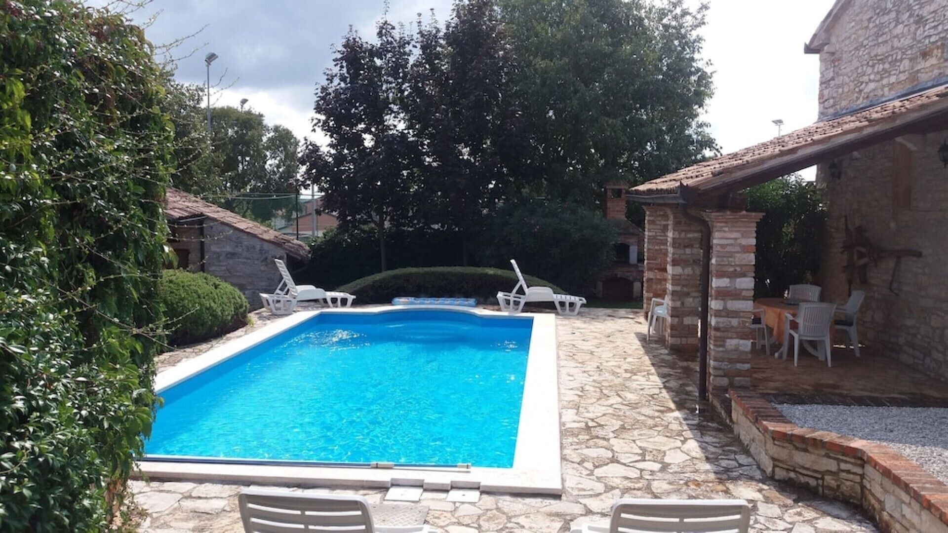 A rectangular swimming pool with a stone deck surrounding it, two white lounge chairs, and a stone building with a tiled roof.