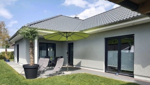 Ein modernes Haus mit einer überdachten Terrasse, einem Rasen und einem großen Sonnenschirm.