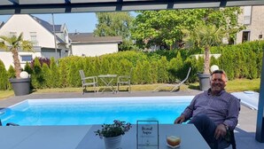 Ein Mann sitzt an einem Tisch im Freien, im Hintergrund ist ein Schwimmbecken zu sehen.