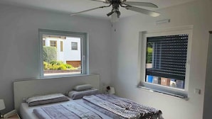 Ein Schlafzimmer mit einem Bett, einem Nachttisch, einem Deckenventilator und zwei Fenstern.