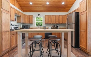 A kitchen with wooden cabinets, a central island with stools, a refrigerator, and a window with a view.