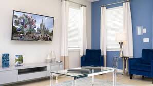 Una sala moderna con una mesa de centro de vidrio, sofás azules y un televisor de pantalla plana mostrando una escena de castillo.